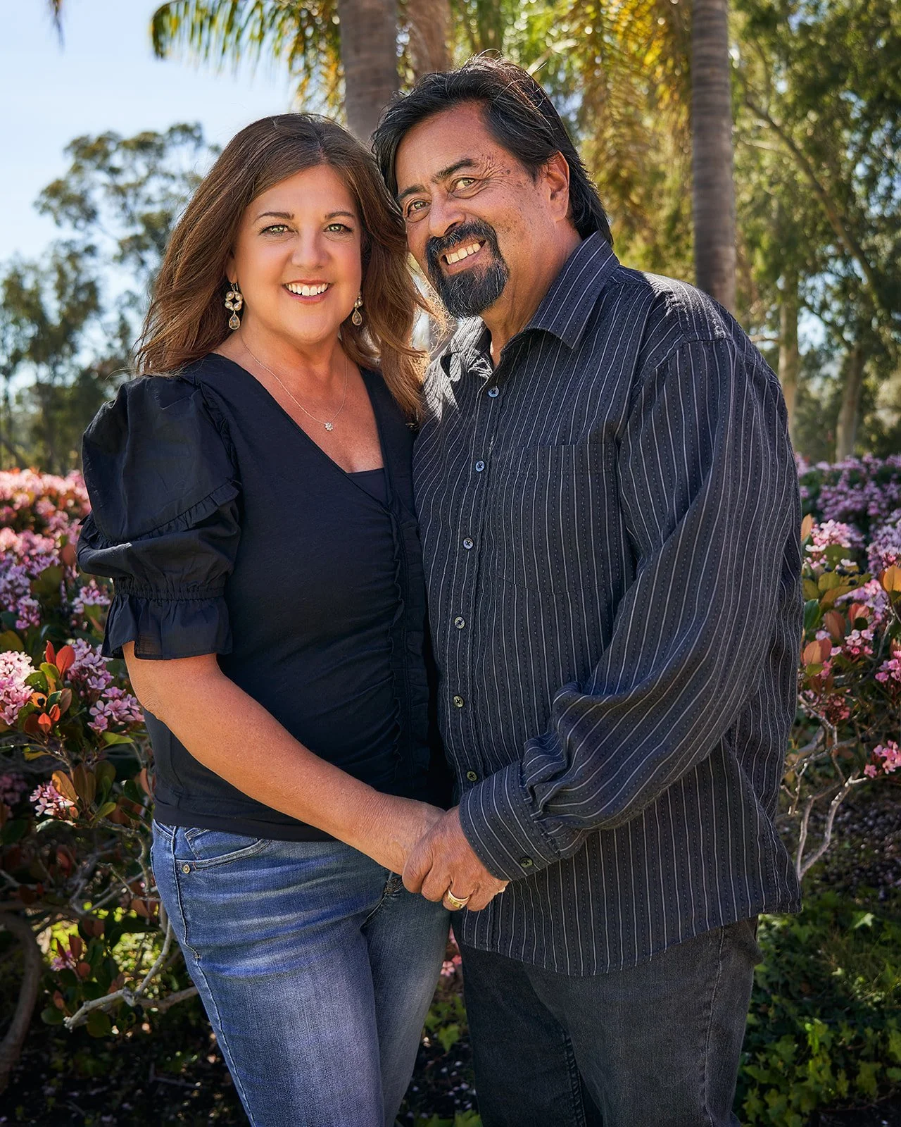 Maria and Rick Sainz smiling together in a garden with pink blossoms behind them.